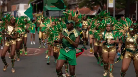 Andrew Aitch/Manchester Carnival A teenage boy wearing a green top and shorts leads a parade of female dancers in gold outfits. They all wear large head-dresses of green and gold feathers.