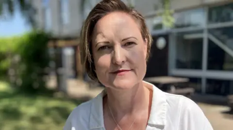 Alison Hernandez is a woman with short brown hair. She is wearing a white blouse and is standing outdoors, near a building, with greenery and picnic benches visible in the background.