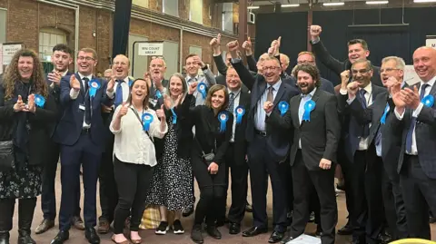 A group of people all cheering in a row for a photo, all Conservatives, wearing blue rosettes, dressed smartly.