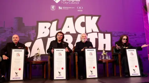 Birmingham City Council From left to right, Bill Ward, Tony Iommi, Geezer Butler and Ozzy Osbourne receive their scrolls. There is a council branded Black Sabbath board behind them in purple. They are sat in front of it on chairs. All are wearing black. Ozzy has his arms outstretched. 