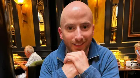 A smiling man sits with his hands below his chin and a blue jumper 