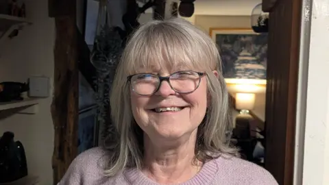 Jacky Smith smiles at the camera inside her home. She has short grey hair and wears glasses along with a light pink knitted jumper.