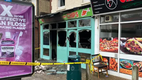 The badly-damaged front of a fish and ship shop in Norwich. The windows are smashed and a yellow piece of tape cordons off the scene.  