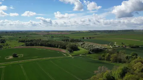 Andrew Chandler An aerial view of farmland showing large green fields, hedgerows, trees, and a small cluster of buildings beneath a cloudy sky.