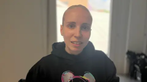 A lady with blonde hair, which has been tied back, wearing a black hooded jumper, stands indoors and smiles at the camera.