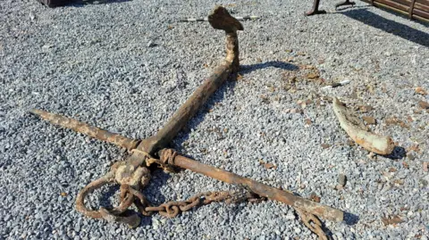 The anchor photographed on land on pebbles. One of its arms appears to be missing. It appears to be near a bench. It's a sunny day.