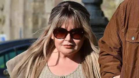 A closeup of a woman entering court. She has dyed blonde hair with darker roots, large sunglasses, and is wearing a cream coloured top.