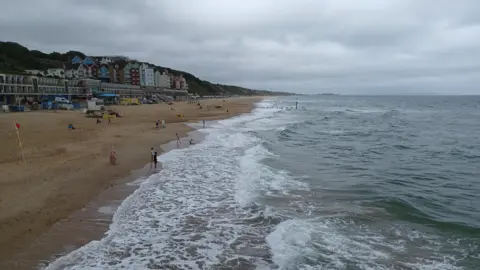 NickyN Boscombe beach on an overcast day. There are a few people on the beach with some children paddling. There are waves breaking onto the beach with white foam and the sky overhead is grey. Behind the beach there are several buildings in front of the cliff and there is a lifeguard station on the beach. 