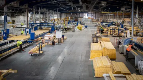 FastHouse An interior of a factory, showing wood stacked into piles. There are several workers in hi-viz jackets and vests working on machinery.