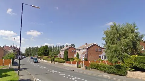A quiet suburban street lined with detached and semi‑detached brick houses. On the right side of the road, the homes have tidy front gardens with bushes, hedges, and a few medium‑sized trees. A low brick wall runs along part of the pavement. On the left side, there is a grassy area with a large tree and a tall wooden fence behind it. Several cars are parked or driving along the road, which has double white lines and a turning lane marked in the centre. Tall streetlights line both sides of the street.