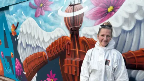 Volunteer Annette standing in front of a mural featuring the Angel of the North appearing though clouds, surrounded by pink flowers. She is wearing white protective overalls. She has dark blonde hair in a ponytail and has sunglasses perched on top of her head.