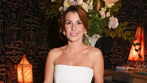 Dave Benett/Getty Images for Disney+ Coleen Rooney wearing a white dress standing in front of a bunch of flowers in a room with black and gold patterned wallpaper