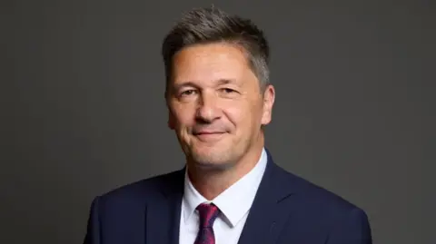 UK Parliament MP John Whitby. He is wearing a blue suit jacket and a dark red tie tinged with a navy pattern.