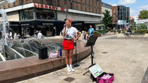 A woman with pink hair is wearing a white t-shirt and red shorts. She is singing out of a microphone and is standing in front of a fountain. 