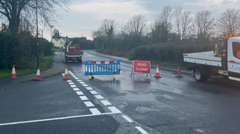 A sign advising that the road is closed on Pagham Road, near Nyetimber, West Sussex, where a car crashed into a gas substation. Several cones are set out on the road.
