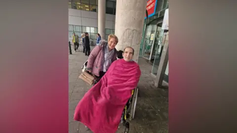 Marshall Wylie Marshall is in a wheelchair and is wrapped a pink blanket. He is outside a hospital and his wife is standing behind him. 