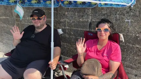 A picture of two people sat on a beach waving at the camera.