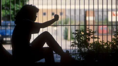 The silhouette of a girl sat crouched on a wall holding onto the metal fence between her and a building. 