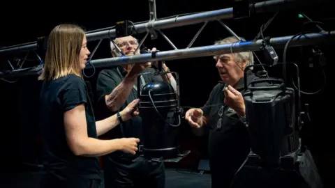 Three people are stood around theatre stage lighting hanging from a rail.
