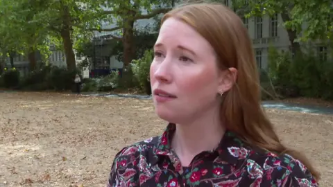 Helen Dollimore stands outdoors in a tree-lined area wearing a patterned dark blouse. Buildings and greenery can be seen in the background.