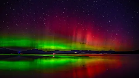 Gerry Doherty Stunning Aurora in shades of red and green light up the sky, reflecting on the water of Loch Lomond, the hills in the distance in silhouette
