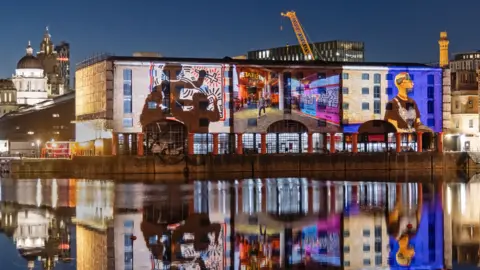 Adam Kenrick Huge projected images are reflected onto a brick building on the water. The images reflect back into the water in the evening's darkness. The projected pictures show Keith Haring cartoon artwork, a picture of the outside of the Tate museum, and a man looking away from the camera.