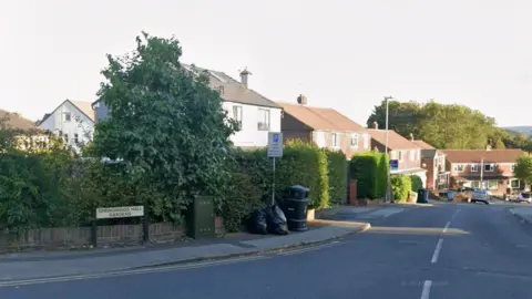 Google Residential street with semi-detached houses featuring red-tiled roofs and white walls. A large leafy tree and trimmed hedges line the pavement. There is a street sign reading ‘Springwood Hall Gardens’ on the left, along with a parking restriction sign and two black rubbish bags near a bin.