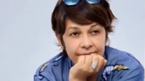A head shot of Karine Buisset, who has her hand on her mouth. She is wearing a blue top and has medium-length brown hair with sunglasses on her head.