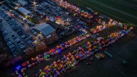 Carl Humphrey A drone image of the festive tractors all lit up and parked in a large car park. Other vehicles are parked nearby without lights. Various buildings can be seen in the middle of the site which is surrounded by fields. 