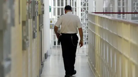 PA Media A general view inside a prison of the back of a prison officer, in a white shirt and black trousers, walking down a prison corridor