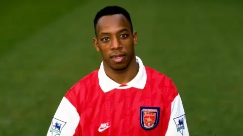 Getty Images An official shot from 1994 of Ian Wright in the Arsenal home strip of the time. It is red with white sleeves, each with a Premier League crest emblazoned on it.