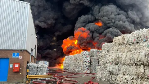 Northamptonshire Fire and Rescue Service Flames and a black plume of smoke are shown rising from a recycling facility. In the foreground, a warehouse building is shown alongside bales of what appears to be plastic-based material.