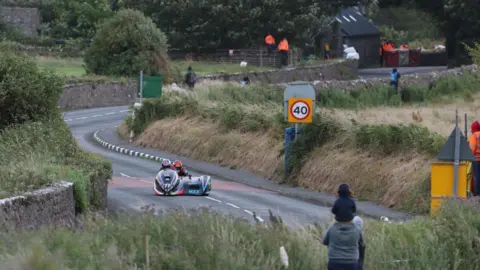 A sidecar on the course during the Southern 100 with the road winding round a corner. A spectator looks on.