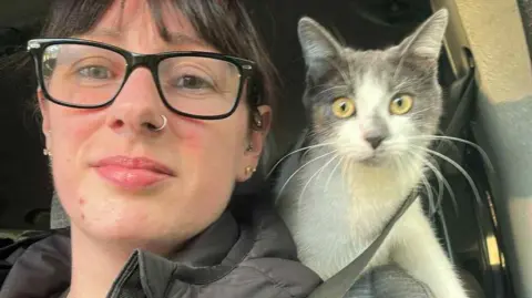Liz Skillicorn Liz Skillicorn and Dusty sit in a car. Dusty looks over her shoulder, liz wears dark glasses, a dark coat and has dark hair. She is also wearing a seat belt.