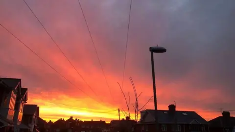 A street lamp is in front of a red sky. The roofs of houses and phone lines can also be seen against the sunset.