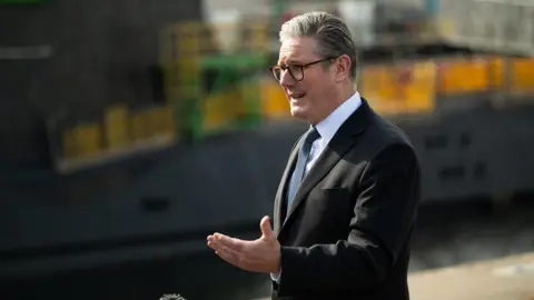 Three quarter profile view of the Prime Minister. He has short greying hair and is wearing spectacles and a dark suit, white shirt and blue spotted tie. He is gesturing with his left hand, with his arm bent at the elbow. The background is out of focus - a blur of grey and yellow.