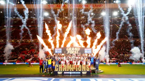 Hull KR celebrate winning the Super League Grand Final - the team all cheer in front of the stands while holding up the trophy as fireworks go off in the background.