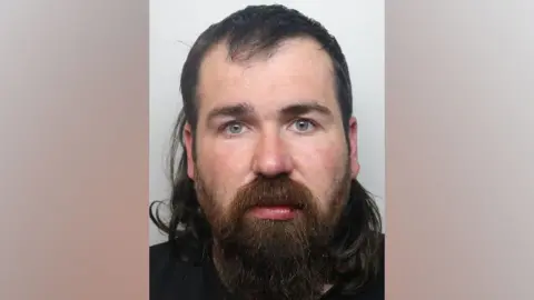 A police mug shot of a man in a black hoodie looking into the camera. He has a long thick beard and long brown hair. 