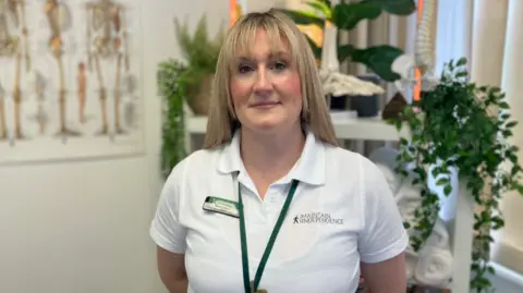 BBC A woman with blonde hair smiles at the camera. She is wearing a white polo shirt with a name badge and a green lanyard around her neck. Behind her is a shelf with plants on it