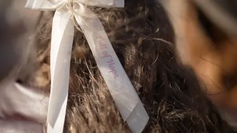 The back of a girl's head. She has brunette hair with a cream hair bow that has the words 'Full of hope' on it.