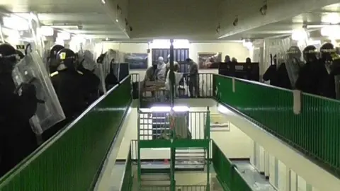 Cambridgeshire Police Lots of police officers with riot equipment are standing either side of a prison wing. In the middle are a group of prisoners who have barricaded themselves into part of the wing. 