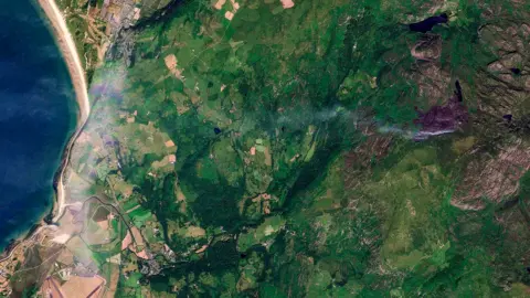 Planet Labs PBC A satellite image of the Rhinogydd mountains in Gwynedd, north Wales, shows plumes of smock. The blue sea is captured on the left side of the image, with fields and mountain landscape on the right-hand side. 