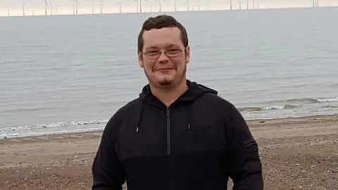 A photo of Aaron Keightley wearing a black hoody. He is standing on a beach with the water behind. 