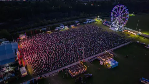 Rich MCD An aerial picture taken at night show's a large crowd in front of the main stage at Good Times live in Chipping Sodbury. In the background of the shot is an illuminated ferris wheel