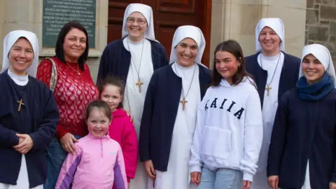 GERRY TEMPLE A group of people including five nuns who are wearing white and blue and have crosses around their necks. The other people are in a woman and three girls - one teenager two younger - standing in casual clothes. They are standing in front of a church.