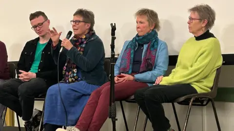 Judy Howard A wide shot of four people sitting in chairs on a platform. On the left is Éanna Mac Cana, who has short brown hair and is wearing a pair of black framed glasses, a black jacket, a green t-shirt and black jeans. On his right is Judy Howard, who is speaking into a wired microphone and has her hand raised. She has short grey hair, a blue jacket, a colourful circle scarf and a long blue skirt. Beside Mrs Howard is her sister. She has short light hair, and is sitting with her phone in her hand looking out at the crowd. She is wearing a patterned scarf, a light blue jacket, a navy t-shirt and maroon trousers. Mrs Howard's second sister is sitting at the end of the row and looking towards the group. She is wearing a pair of thin framed glasses, a black turtleneck and a neon yellow jumper over top with a pair of black trousers.