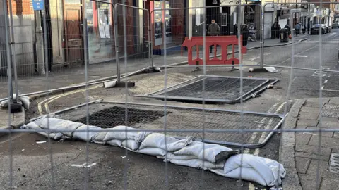 A grey road with two sinkholes and metal grey fencing covering them. 
