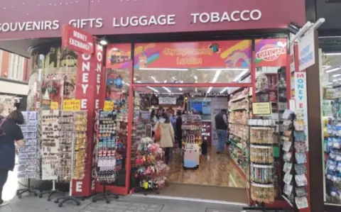 A candy store on Oxford Street. Outside are stands of tourist memorabilia such as magnets and post cards, while inside the shop there are items such as suitcases and sweets