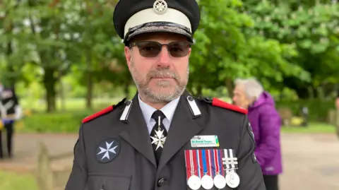 A man dressed in a dark grey St John Ambulance Service uniform is stood outside with trees in the background. He is wearing a black cap with the charity's logo. He has four silver medals on his lapel. He has a grey-coloured beard and moustache. He's wearing sunglasses.