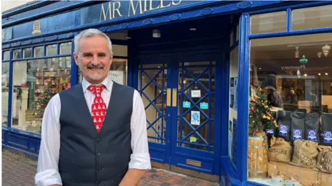 James Briggs Colin Barrell standing outside his tearooms in Taunton smiling at the camera. He is wearing a dark grey waistcoat, white long-sleeved shirt and a red and white patterned tie. He has a closely-cropped beard and moustache and short greying hair